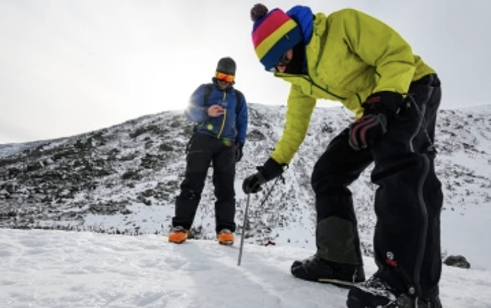 Two people in winter gear are examining the snow on a snowy mountain landscape.